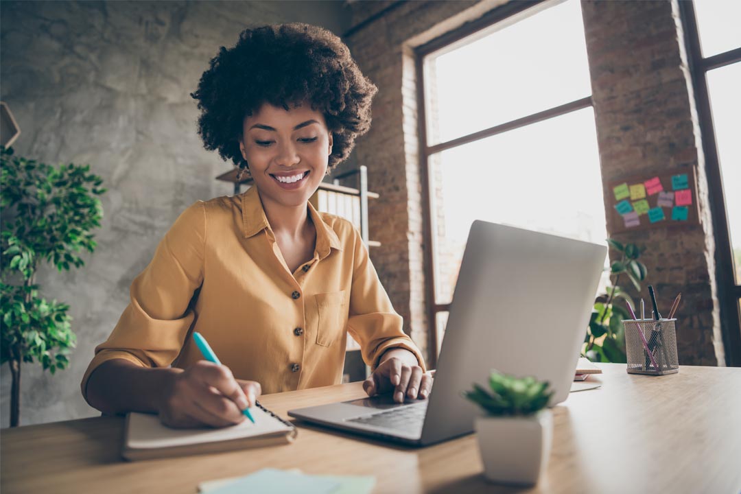 Person smiling and working on laptop with notepad in modern office environment